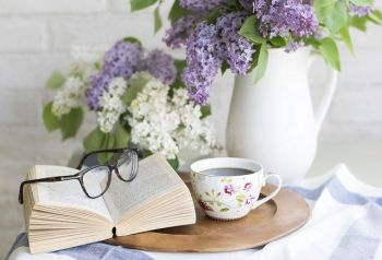 Kahvikuppi puutautasella, kirja vieressä, jonka päällä silmälasit, kannuss sireeninoksia.