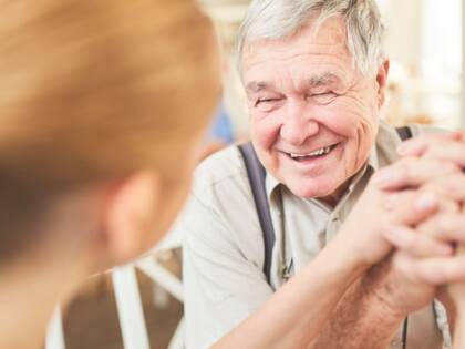 An elderly man is sitting opposite a younger woman. They are holding hands and laughing.