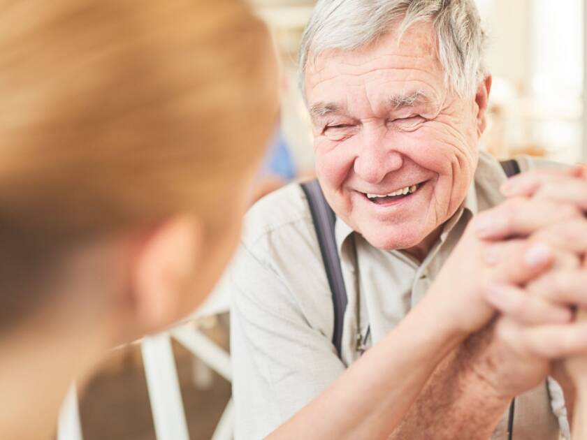 An elderly man is sitting opposite a younger woman. They are holding hands and laughing.