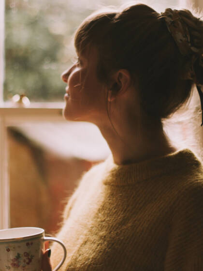 A woman is looking out a window and she is holding a mug in her hand.
