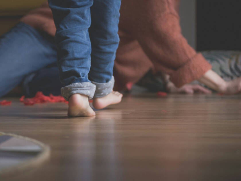A child walks with bare feet on a wooden floor with an adult in the background.