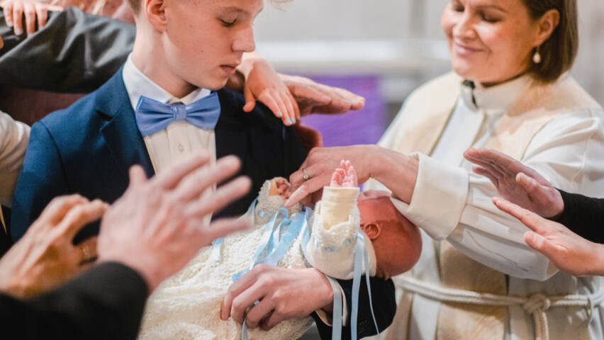 Kastettava vauva on nuoren pojan sylissä, ja pappi ja läheiset ovat nostaneet kätensä siunatakseen vauvaa.