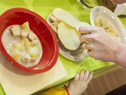 Ruokapöydällä on punaisessa kulhossa kalakeittoa ja valkoisella lautasella leipää, johon lisätään juustosiivuja. Lapsen käsi näkyy alareunassa.