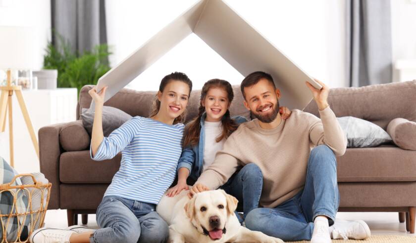 Perhe olohuoneessa lattialla. Mukana on myös kultainen noutaja koira. He ovat astelleet päällensä leikkimielisen kodin katon.