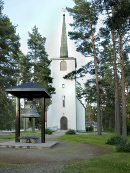 Valkoinen, vaaleaksi rapattu kirkko, jossa on terävä torni, sijaitsee metsän keskellä. Kirkon edessä on suuri kirkonkello katetussa telineessä. Vihreä nurmikko ympäröi kirkkoa.