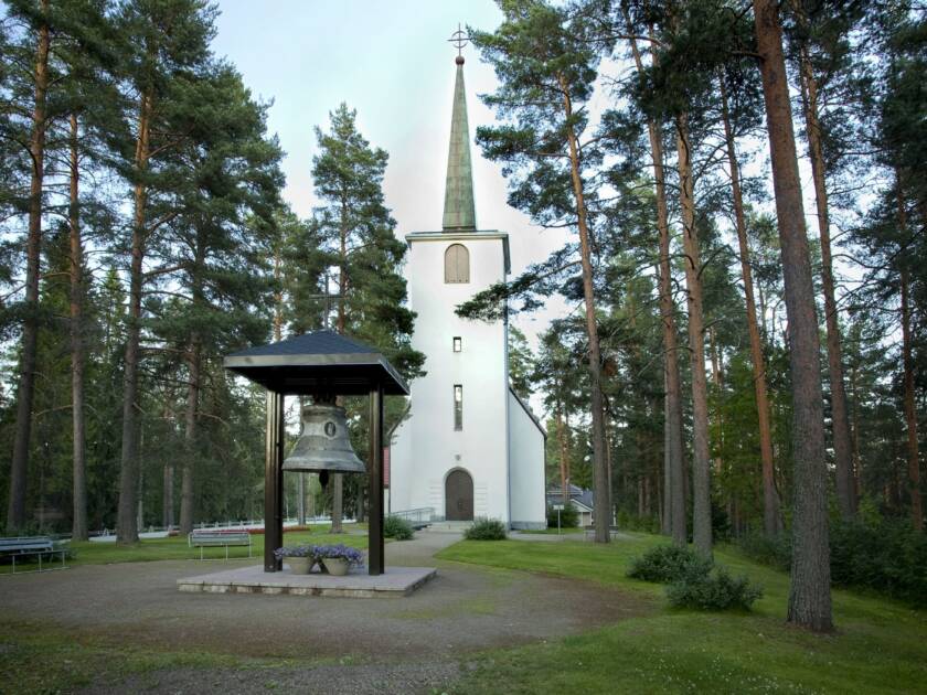Valkoinen, vaaleaksi rapattu kirkko, jossa on terävä torni, sijaitsee metsän keskellä. Kirkon edessä on suuri kirkonkello katetussa telineessä. Vihreä nurmikko ympäröi kirkkoa.