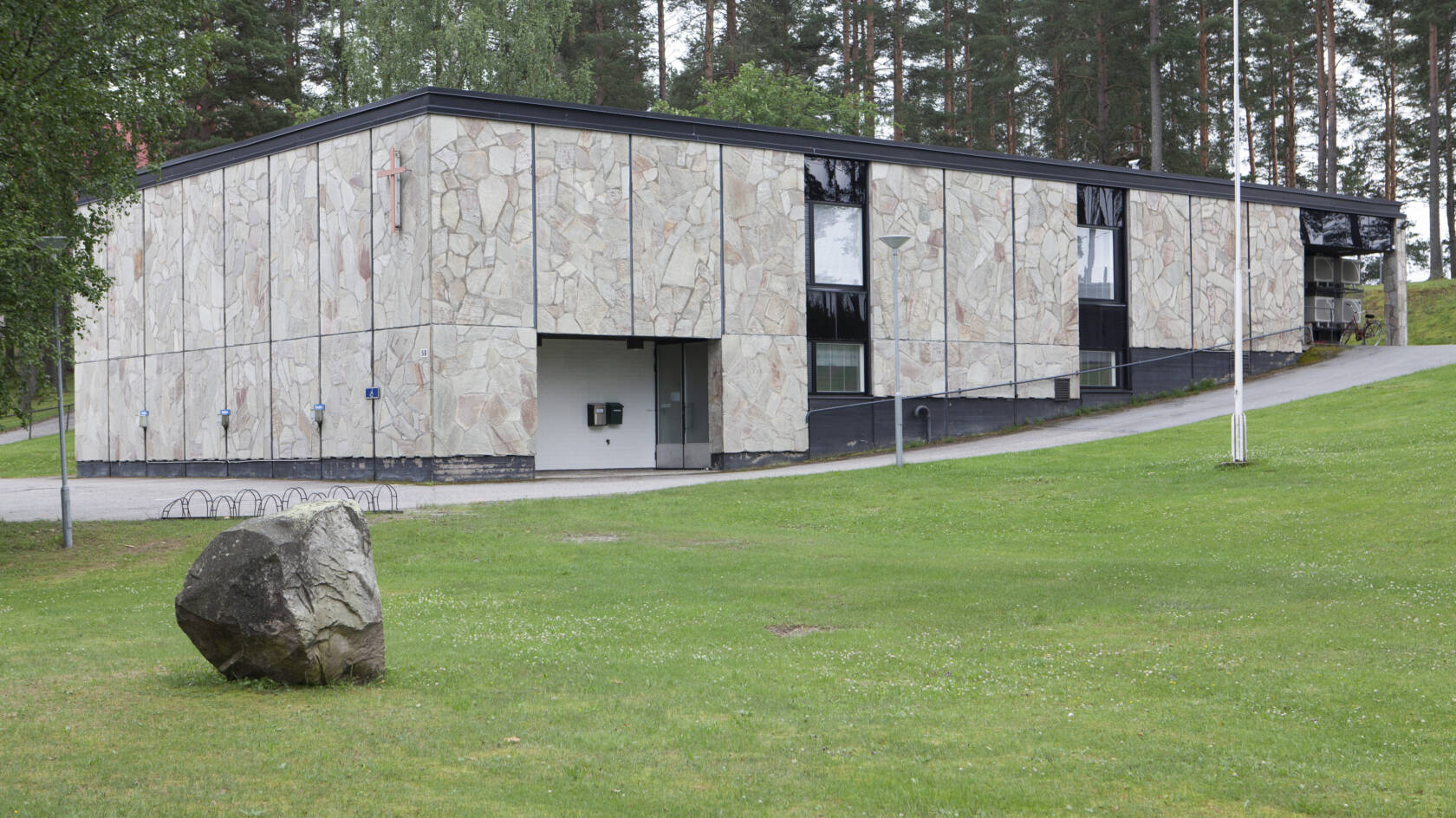 Moderni rakennus, jossa on kivi- ja lasiseiniä, ympäröity vihreällä nurmella ja puilla. Sisäänkäynti näkyy vasemmalla. Nurmikolla on suuri koristekivi.