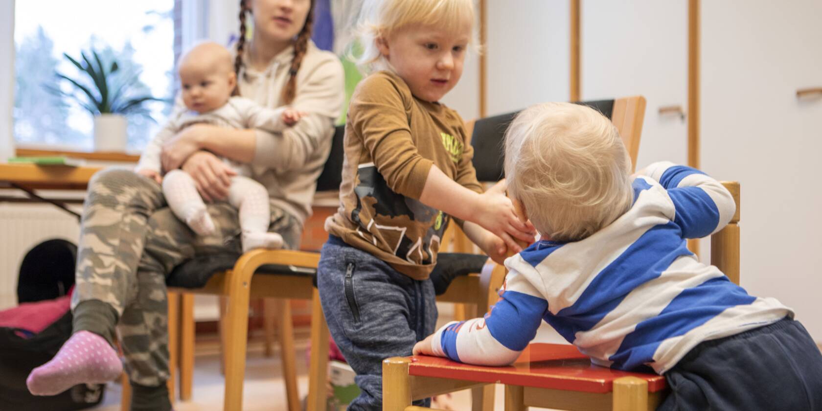 Äiti istuu tuolilla vauva sylissään, kun kaksi lasta leikkii lattialla. Toinen lapsista on tuolilla, toinen seisoo ja pitää kädestä.