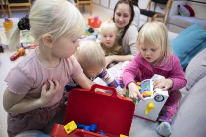 Lapsia leikkimässä lattialla, yksi tutkii punaista lelulaatikkoa, toinen leikkii lelulaitteella, taustalla aikuinen ja muita lapsia.