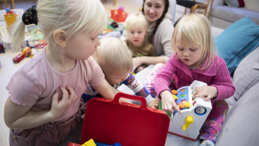 Lapsia leikkimässä lattialla, yksi tutkii punaista lelulaatikkoa, toinen leikkii lelulaitteella, taustalla aikuinen ja muita lapsia.