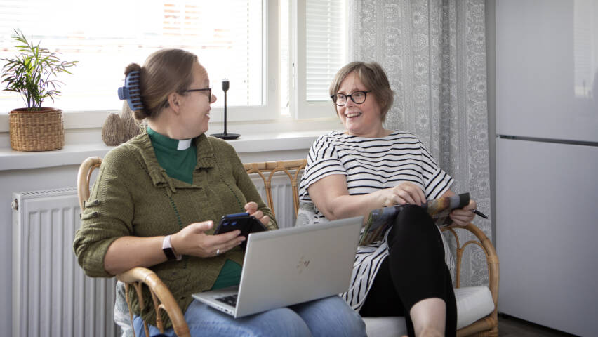 Diakonin kotikäynti. Kaksi henkilöä istuu vierekkäin ikkunan edessä juttelemassa.