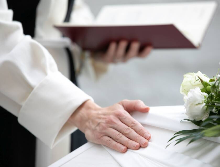 Hautajaisissa pappi pitää kirjaa ja koskettaa arkkuun asetettua valkoista kangasta, taustalla kukkakimppu.