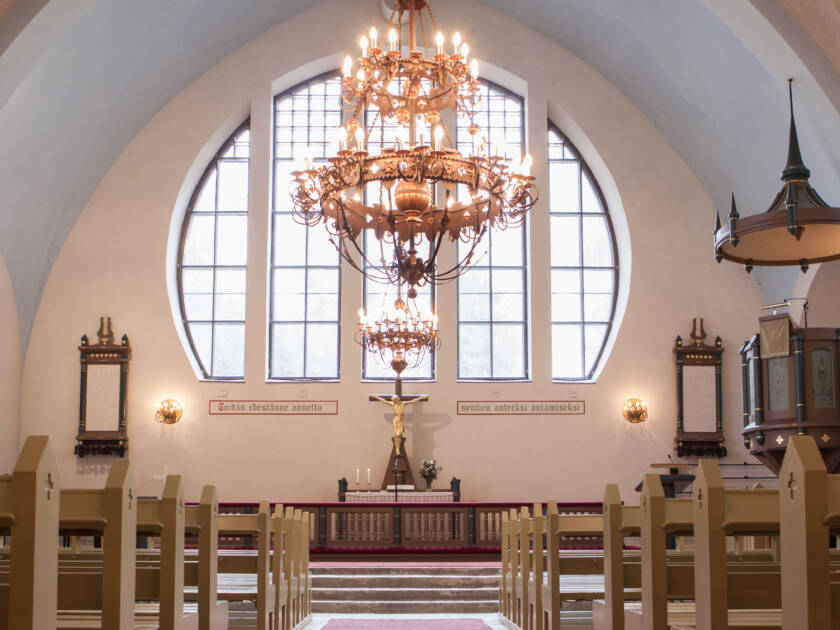 Kirkon sisätila, jossa on suuri ikkuna, kynttilävalaisimia ja alttari. Taustalla näkyy puinen saarnatuoli ja penkit.