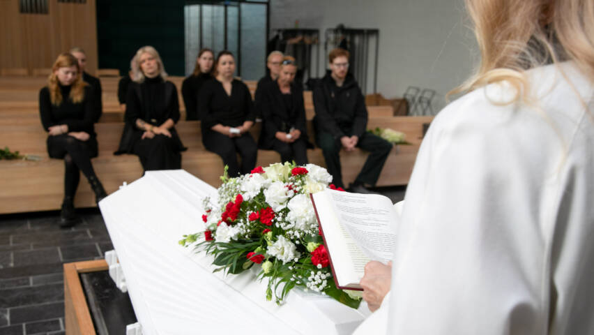 Pappi etualalla arkun vieressä. Taustalla istuu omaisia kappelin penkissä.