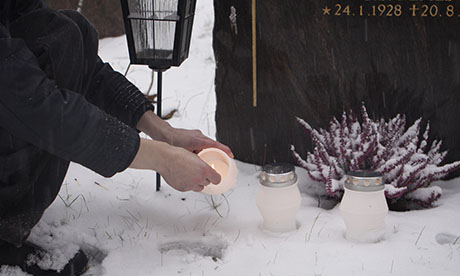 Mies sytyttää kynttilän hautakiven edessä lumisessa ympäristössä, taustalla näkyy hautakivi ja muita kynttilöitä.